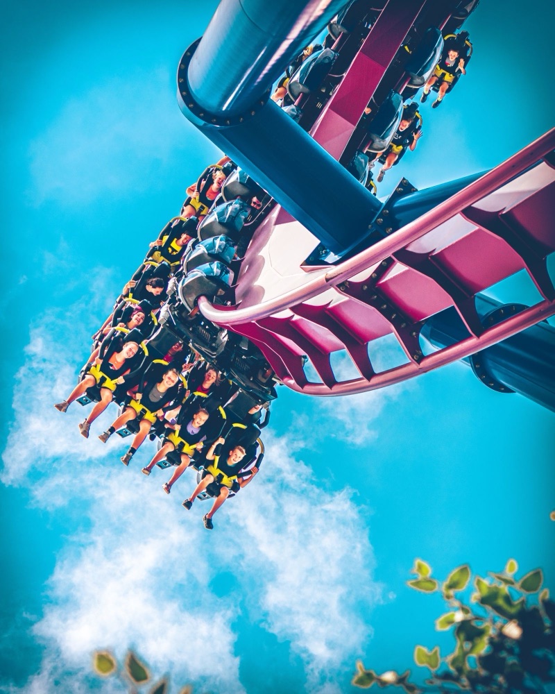Photograph from below: riders with their legs dangling on an inverted coaster train against a bright blue sky.