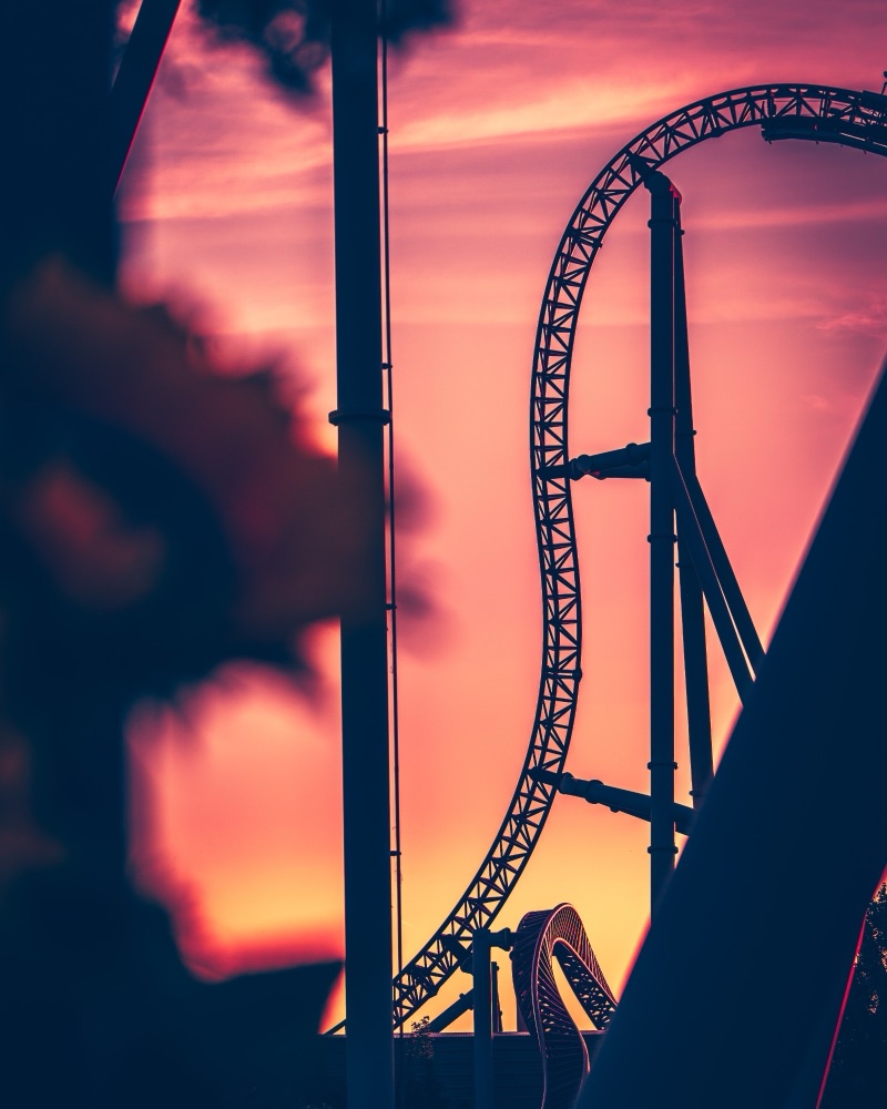 Photograph: roller coaster track and supports silhouetted against a pink and orange sunset.